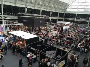 Busy trade show hall seen from a balcony, with rows of exhibitor stands and crowds of visitors under a glass roof