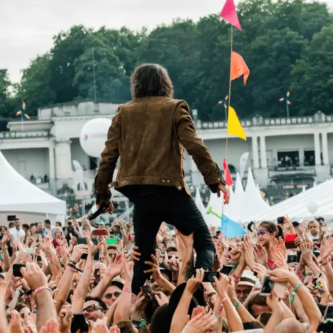 Performer i brun jakke crowdsurfer på udendørs festival, farverige flag over jublende publikum.