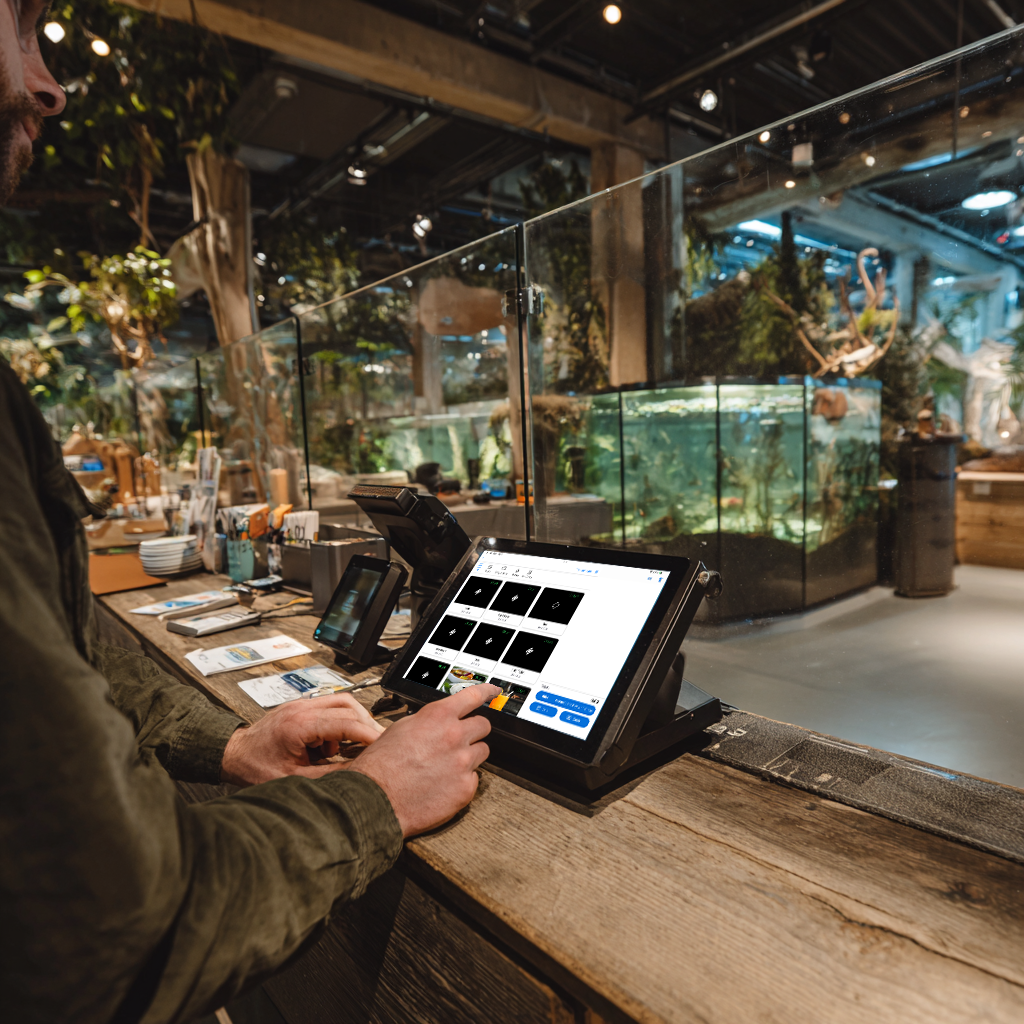 Person, der bruger touchscreen Billetto POS-system ved indgangsdisken i zoo med akvarier og udstillinger i baggrunden